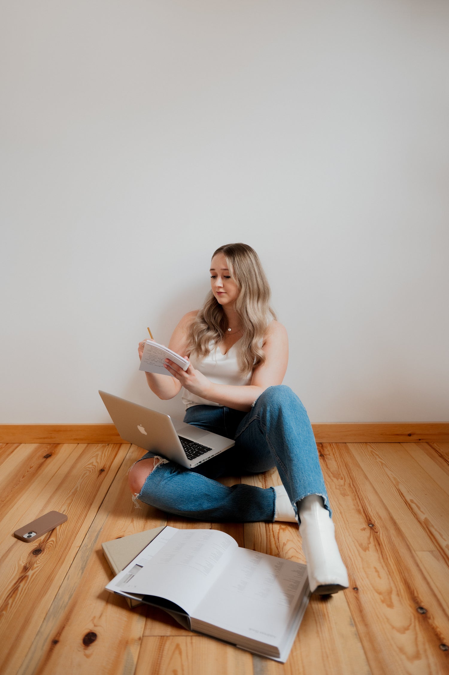 Blonde woman, Emma Curits, sitting on wooden floor with back to wall taking notes regarding marketing strategy for a small business